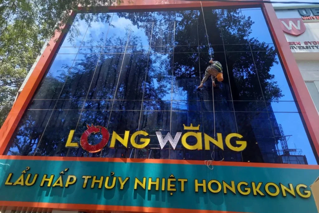 Nhân viên vệ sinh chuyên nghiệp đang lau kính mặt tiền tòa nhà cao tầng bằng phương pháp đu dây, đảm bảo sạch bóng và an toàn.
